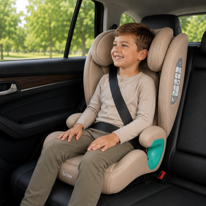 A smiling young boy sits in a beige car seat with a black harness inside a black car, with green trees visible through the window background.