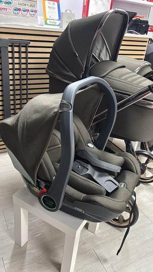 Gray stroller with mounted car seat on white chair display, padded headrest and harness